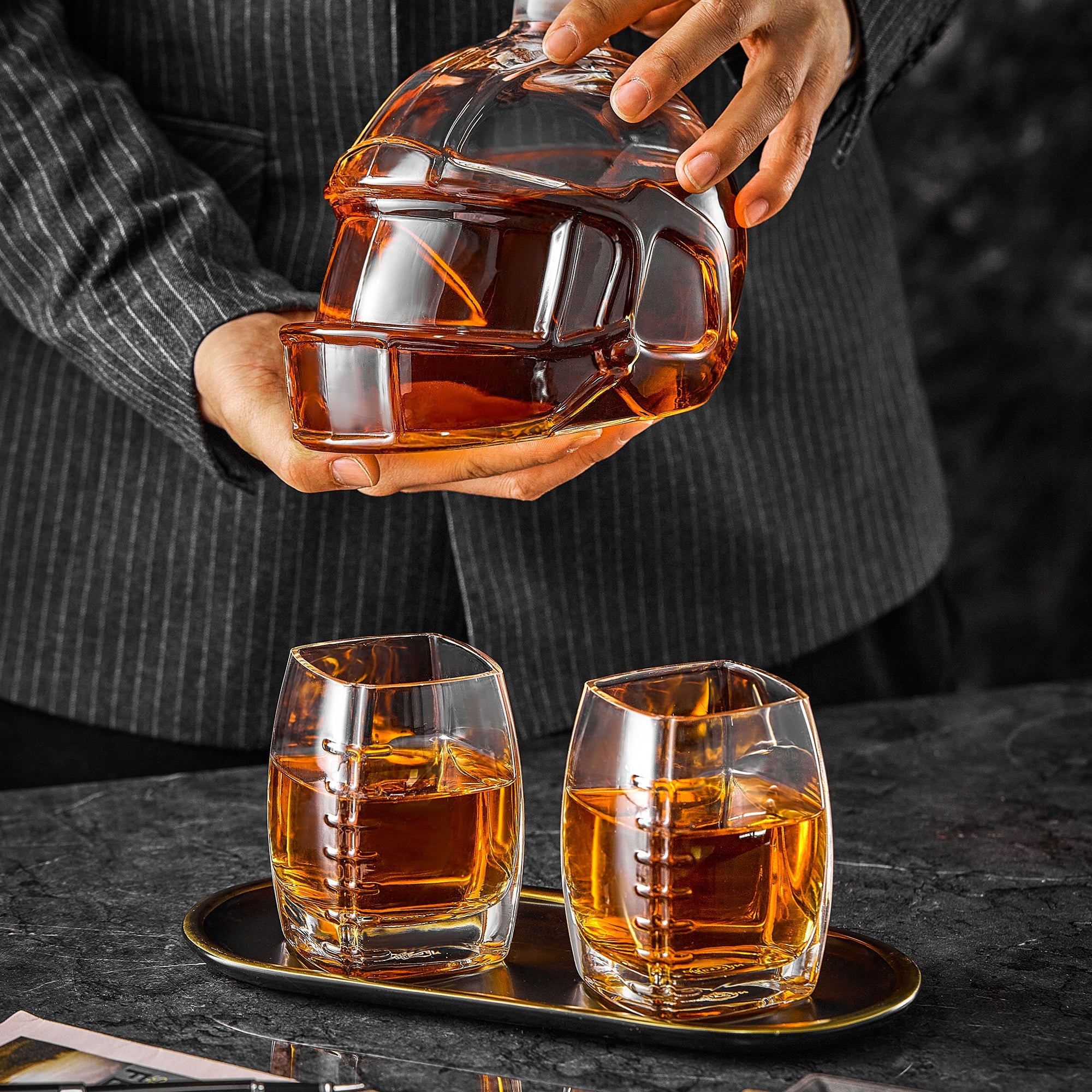Football-shaped whiskey decanter being held above two amber tumblers