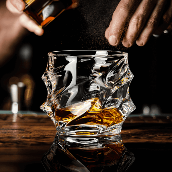 Crystal whisky glass with amber liquid, pour in progress on a wooden surface