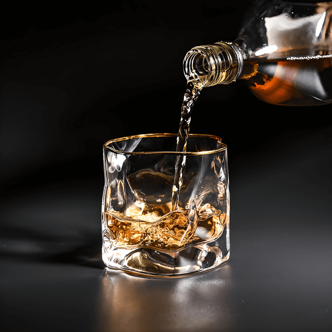 Gold-rim whisky glass with amber liquor being poured into faceted tumbler