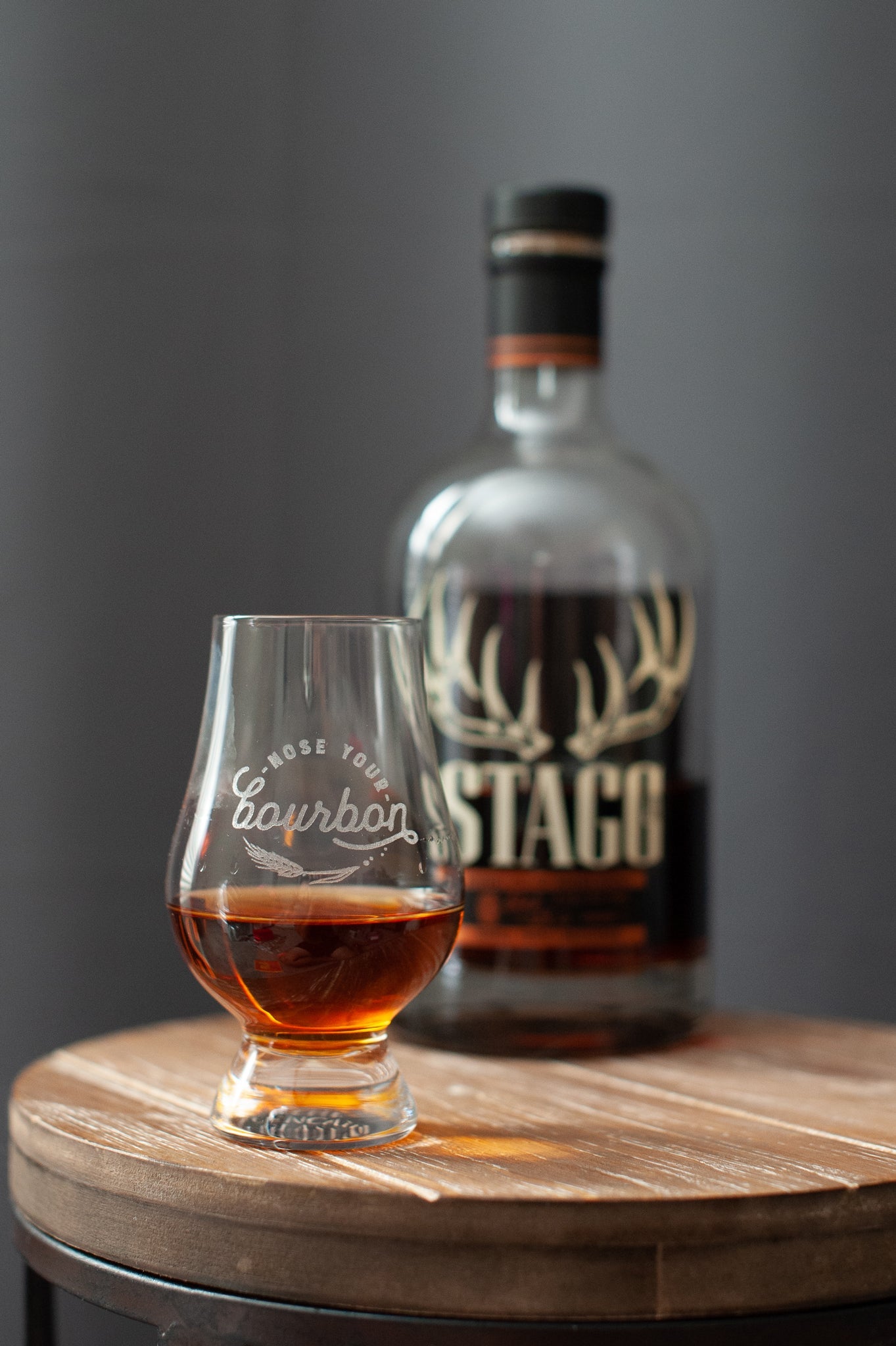 Bourbon tasting glass with amber drink and engraved logo, beside bottle on wood