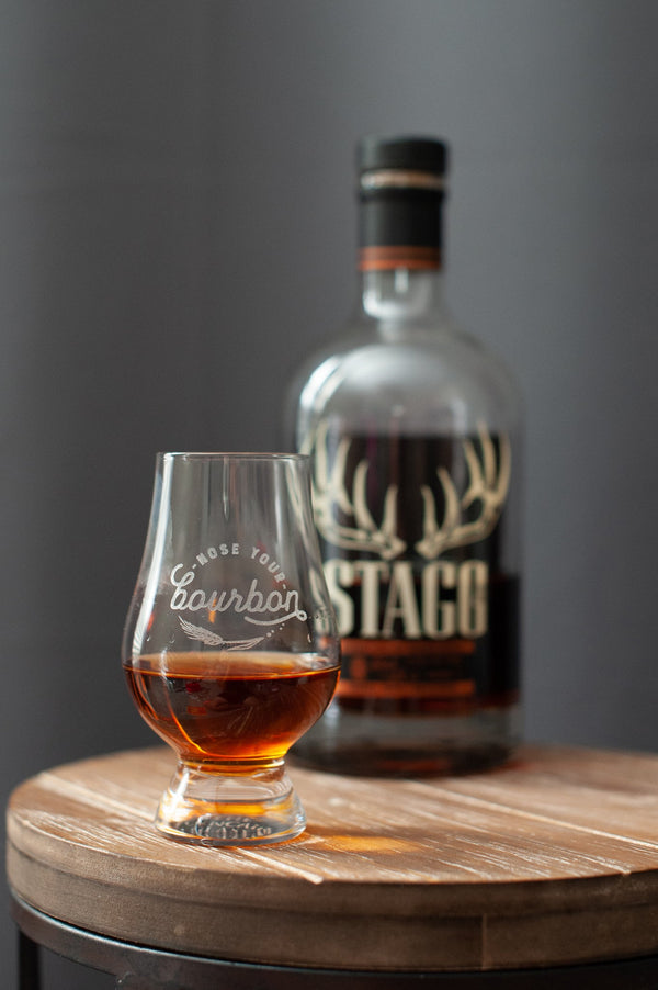 Bourbon tasting glass with amber drink and engraved logo, beside bottle on wood