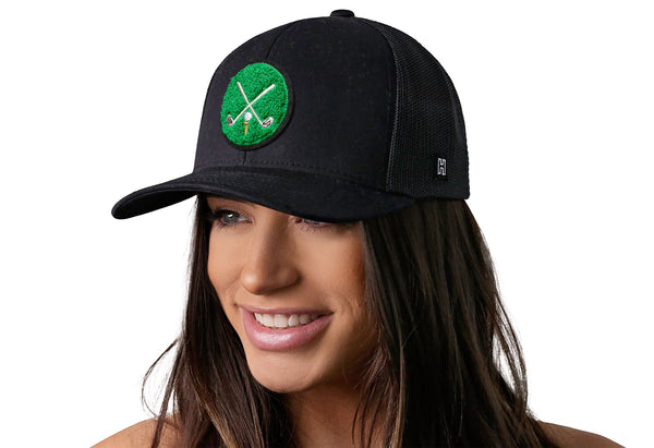 Black trucker cap with green crossed clubs patch worn by woman, front mesh panels.