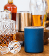 Blue insulated whiskey lowball tumbler on wooden surface with glass bottles nearby