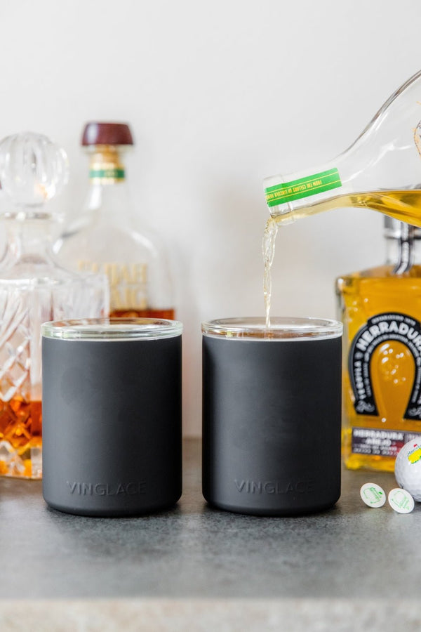Two dark glass-lined lowball whiskey tumblers on a table with liquor bottles