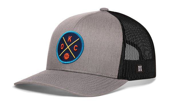 Gray and black trucker hat with mesh sides, curved brim, and circular team logo patch.