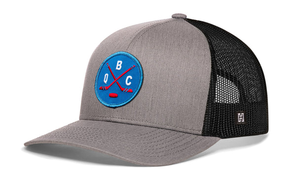 Gray hockey trucker cap with blue front logo patch and black mesh side panels, curved brim