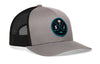 Gray trucker cap with black mesh side panels, circular front logo patch, curved brim.